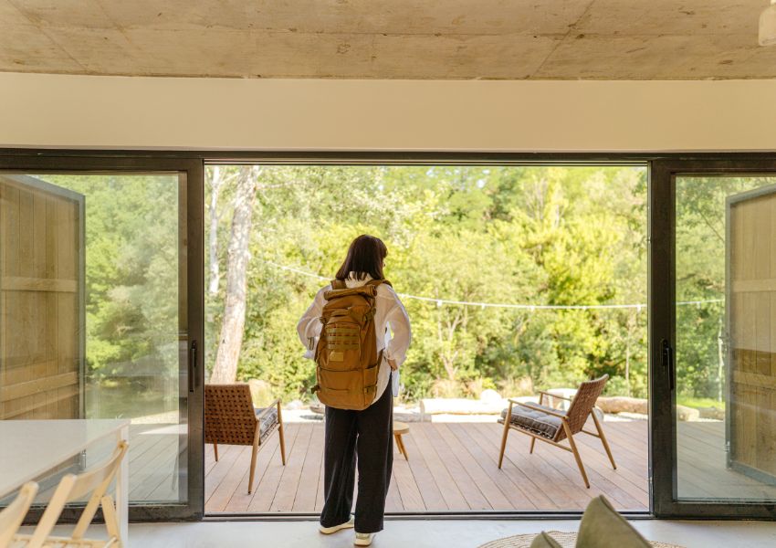 A traveller with a brown backpack stands in the center of a vacation rental home, looking out its vast windows onto the porch surrounded by trees.